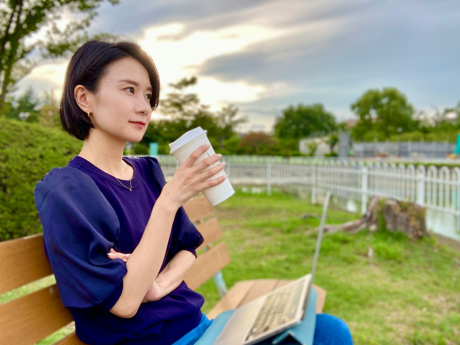 坐在公园长椅上的女性手持咖啡并使用笔记本电脑