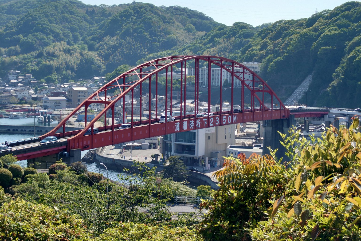 Ponte ad arco rosso che attraversa un fiume con colline verdi e una città sullo sfondo