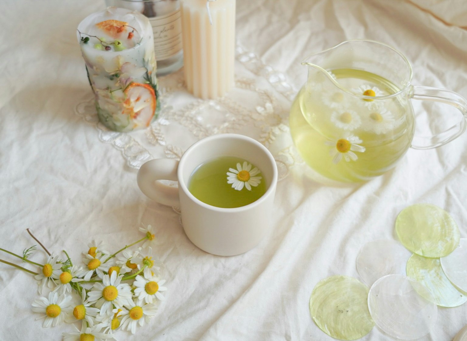 A cup of green tea with a chamomile flower floating on top next to a pitcher of tea and fresh salad rolls on a white cloth