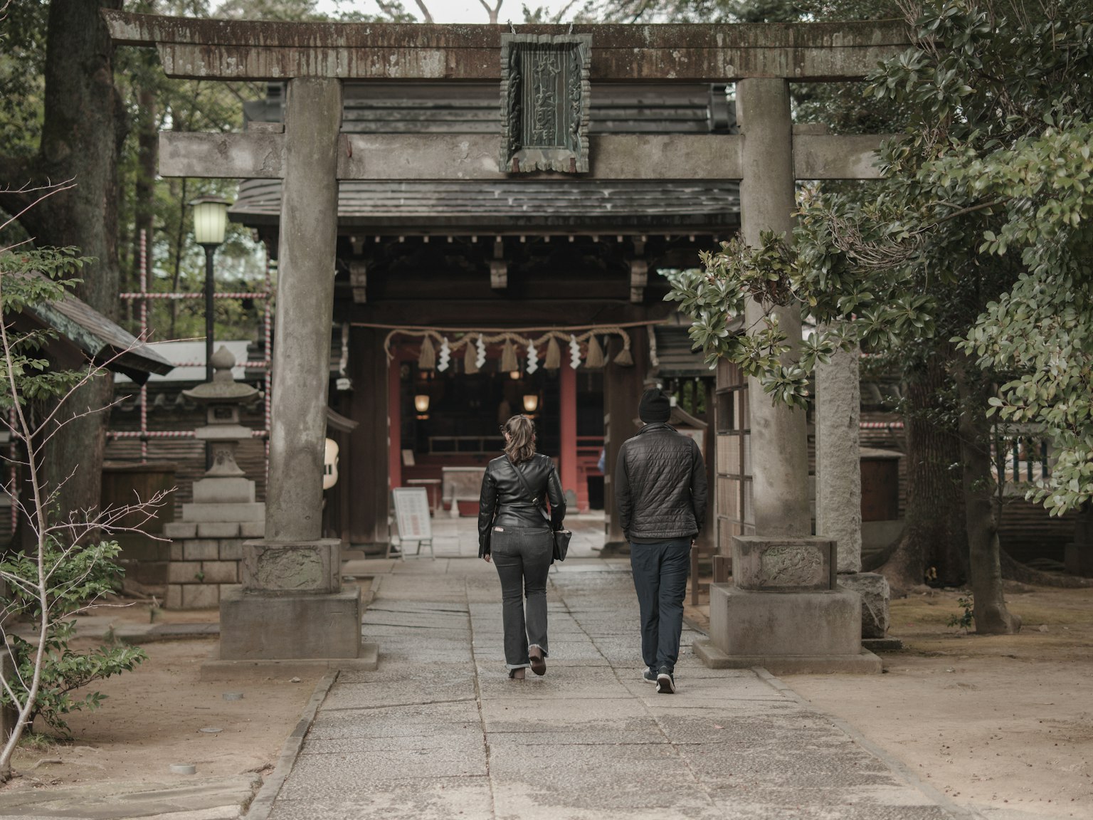 Two people walking towards a shrine with an ancient stone gate and lush greenery