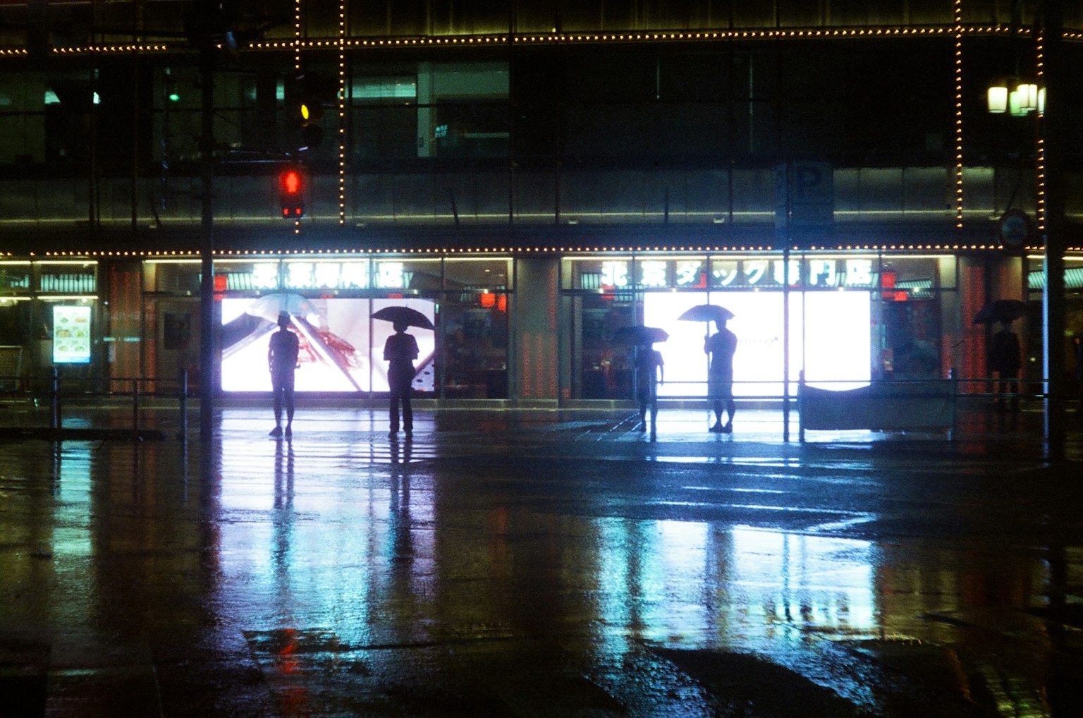 Silhouettes of people holding umbrellas with bright advertisements reflected on a rainy street corner