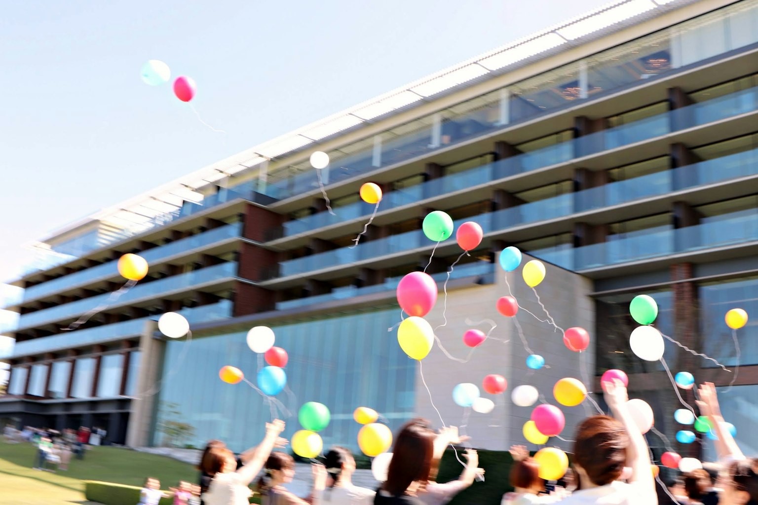 Des personnes lâchant des ballons colorés dans le ciel lors d'un événement avec un bâtiment moderne en arrière-plan