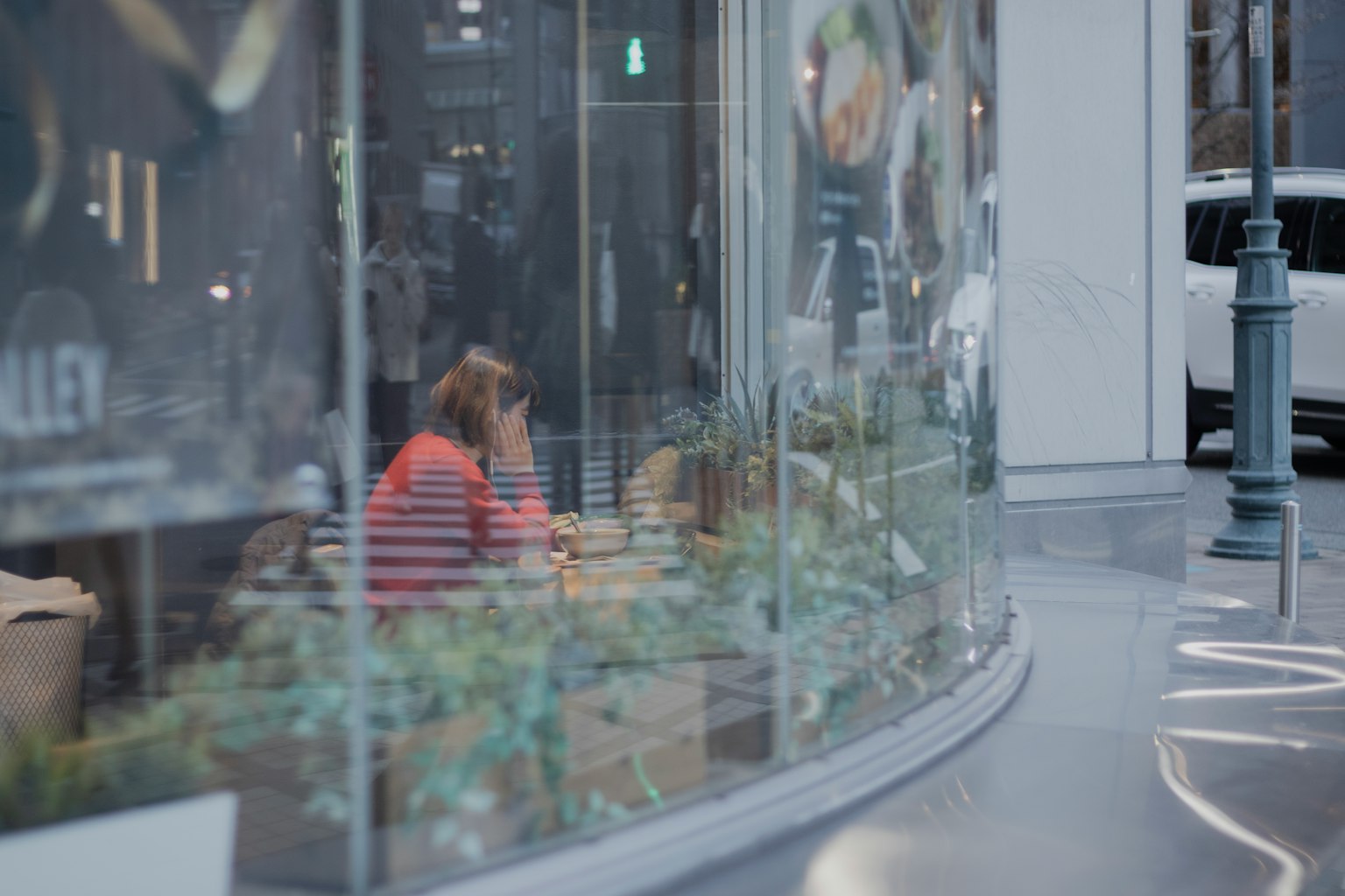 Una donna persa nei suoi pensieri all'interno di un caffè visibile attraverso il vetro con piante circostanti