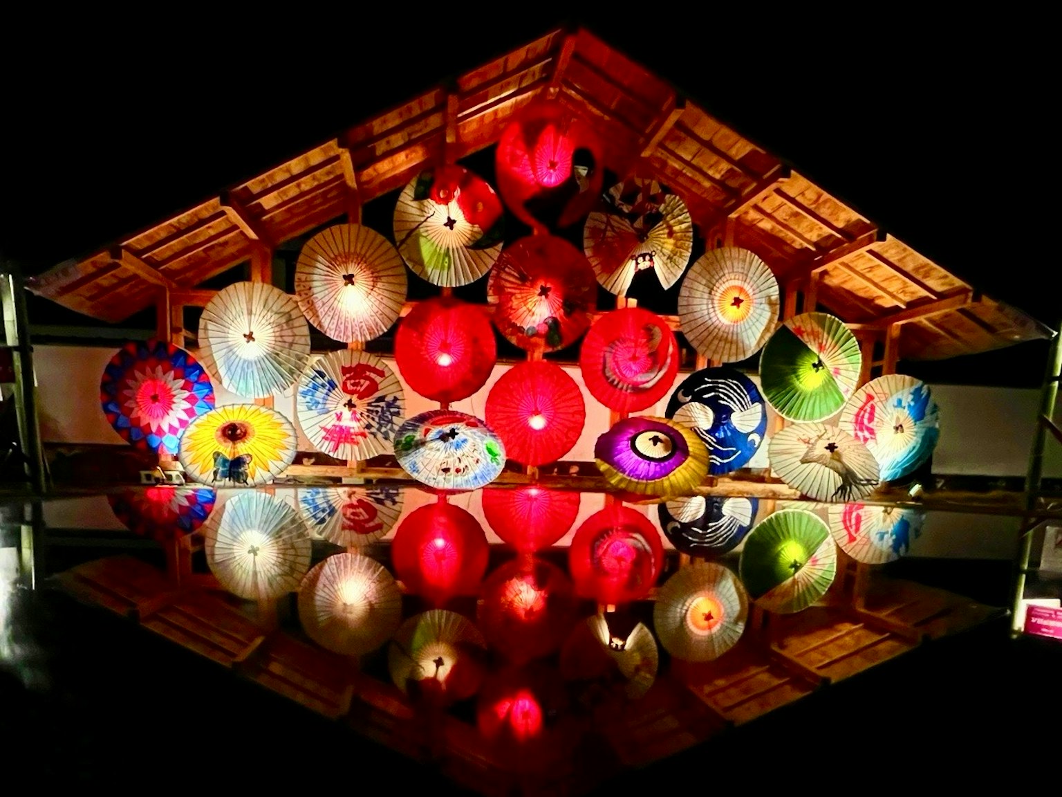 Colorful umbrellas illuminated at night under a wooden roof reflecting on water