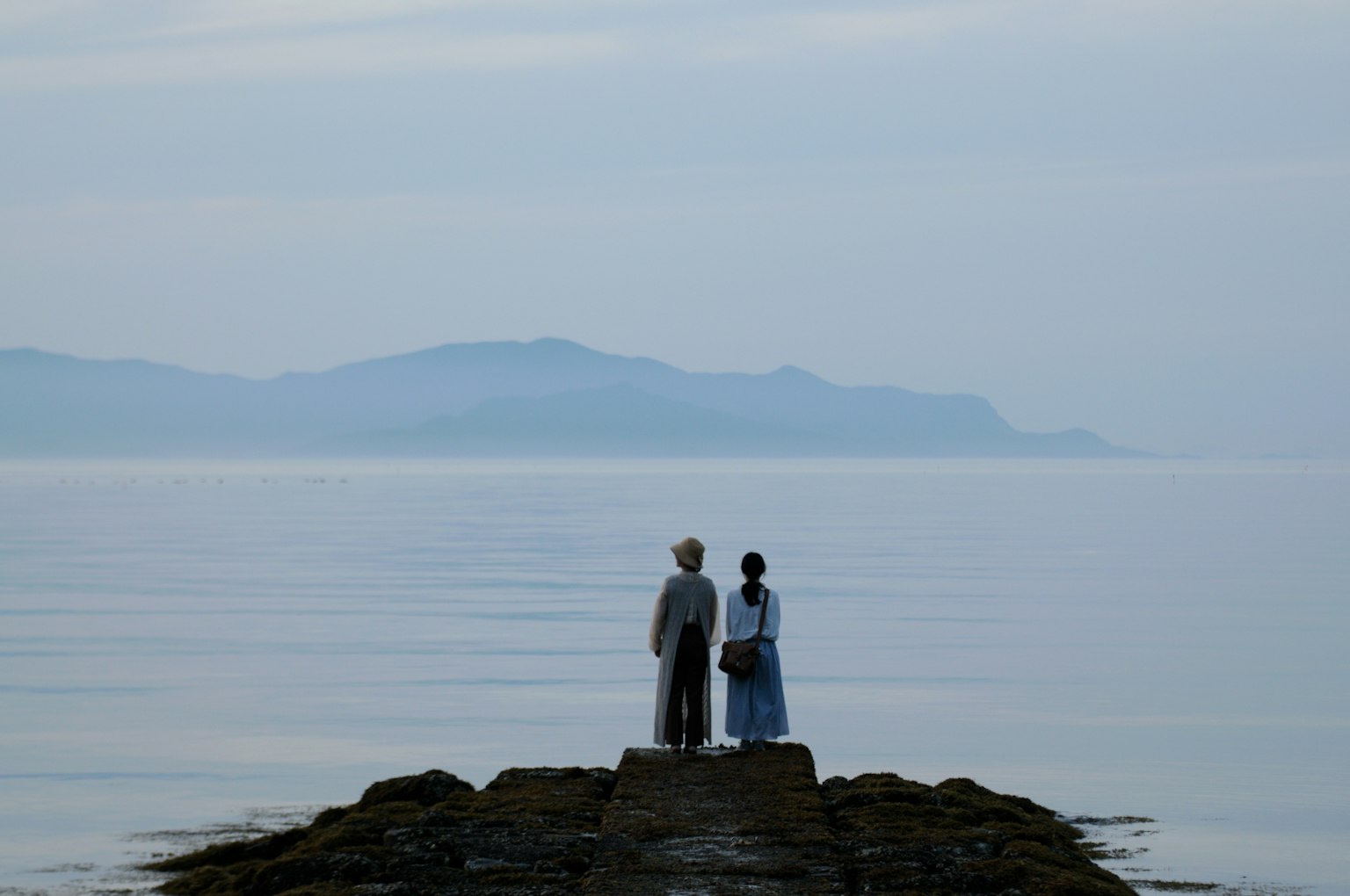 Deux figures se tenant sur un quai rocheux surplombant une mer calme
