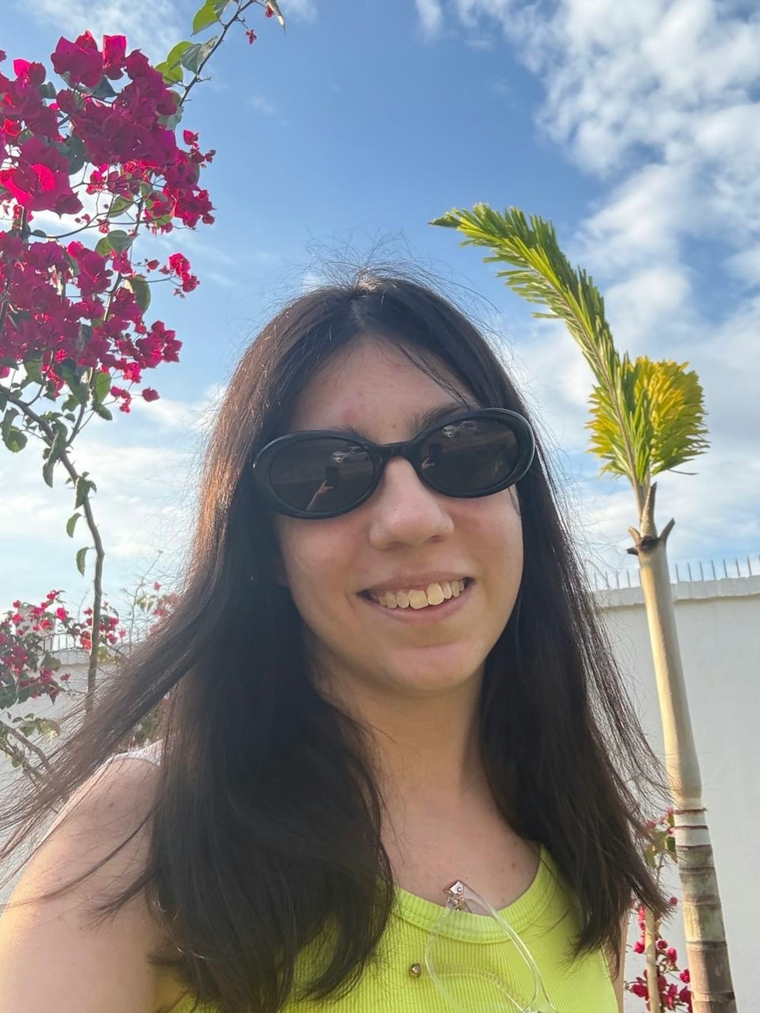 明るい黄色のトップを着た女性がサングラスをかけて微笑んでいる背景にはピンクのブーゲンビリアとヤシの木が見える