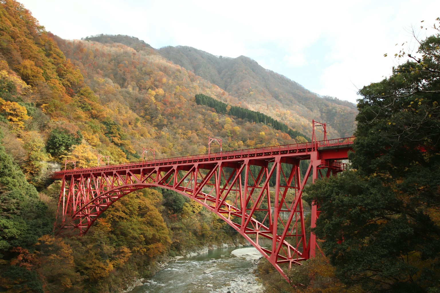 A red iron bridge spans between mountains adorned with autumn foliage