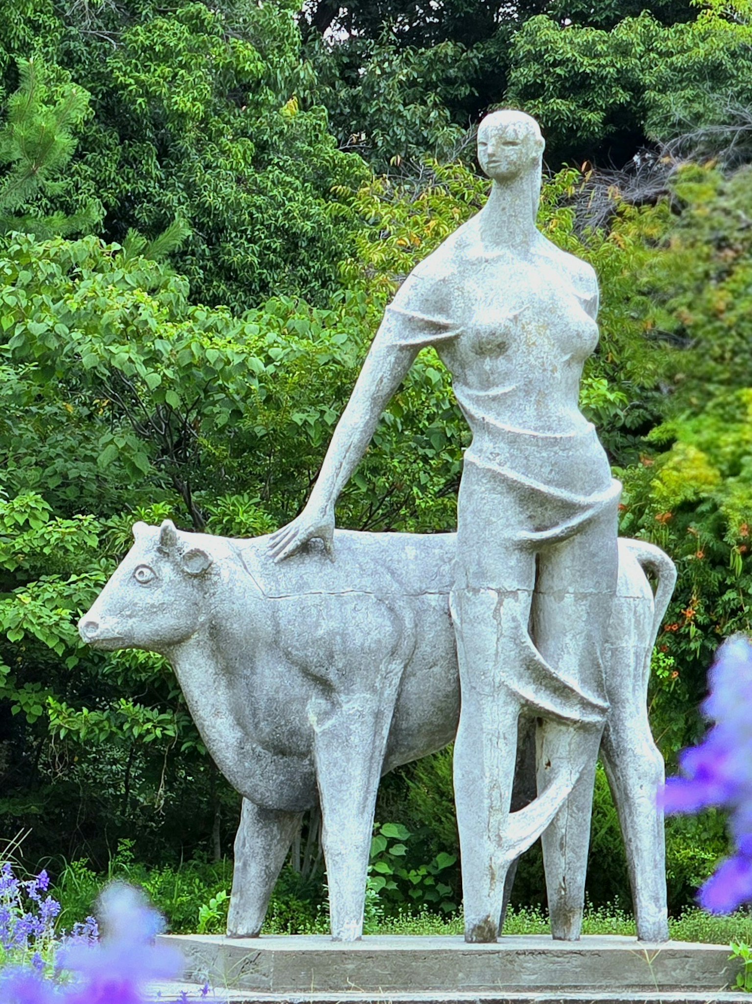 女性の彫刻が牛を撫でている彫刻作品 緑豊かな背景