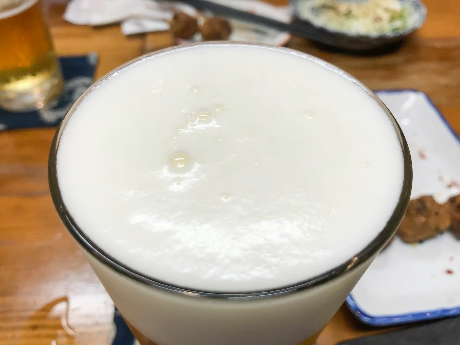 Un bicchiere di birra con una schiuma bianca spessa e cibo visibile sullo sfondo