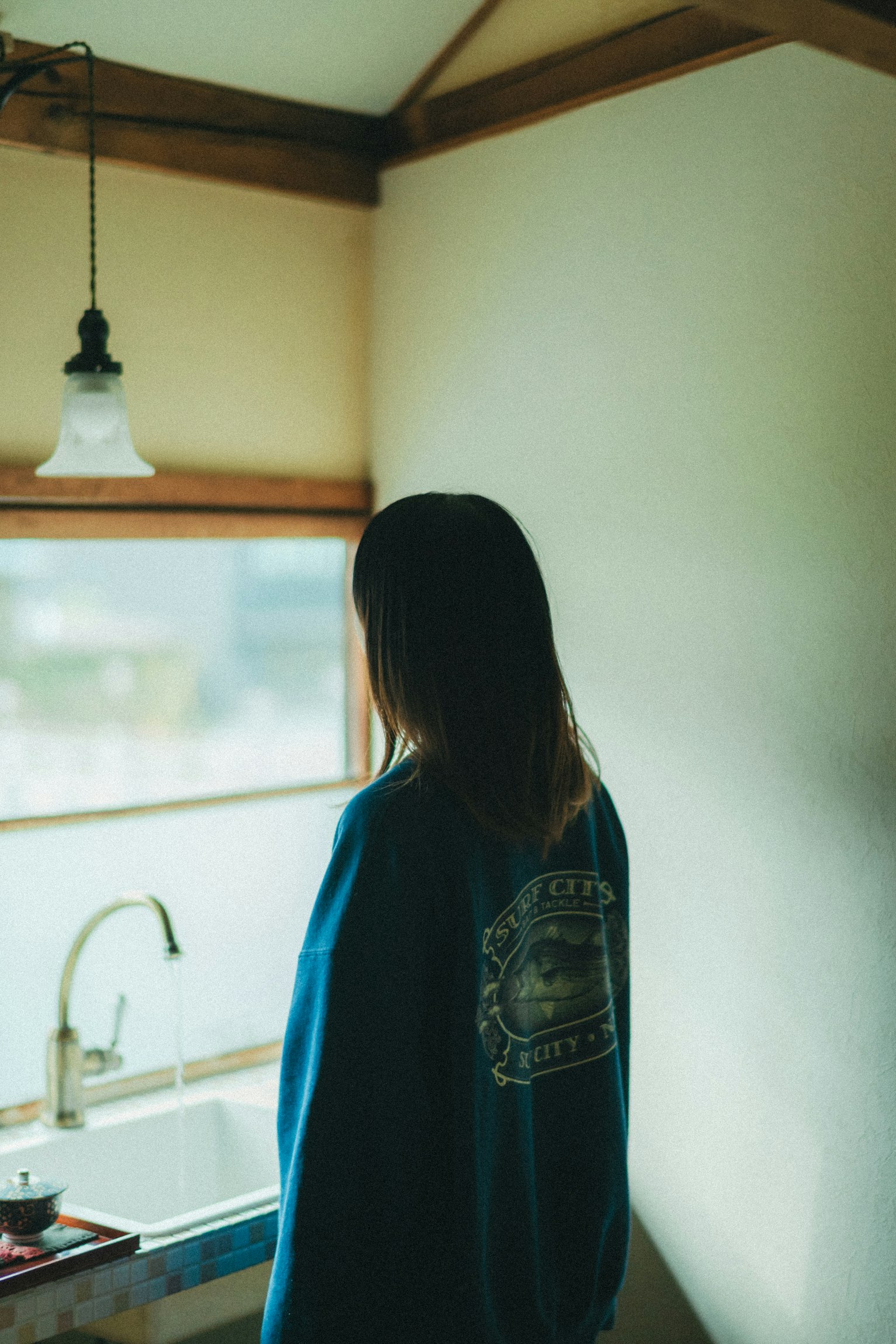 窓の近くに立つ女性の後ろ姿 青いスウェットシャツを着ている 明るい室内の風景