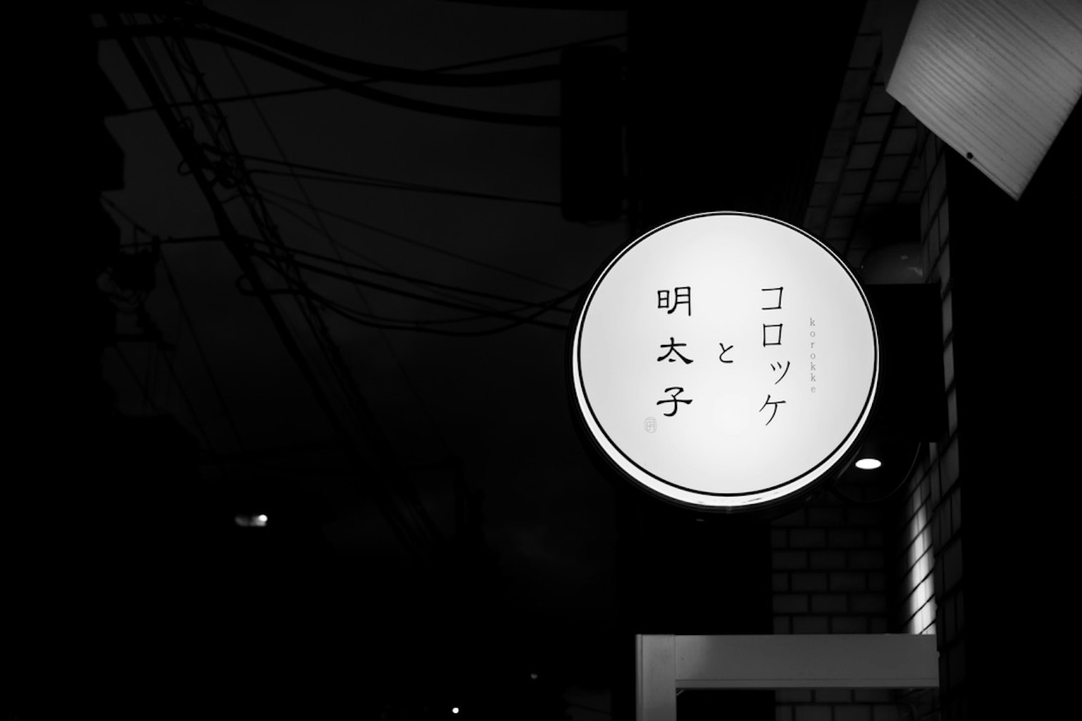 Un letrero redondo blanco con el texto 明太子 y コロッケ sobre un fondo oscuro