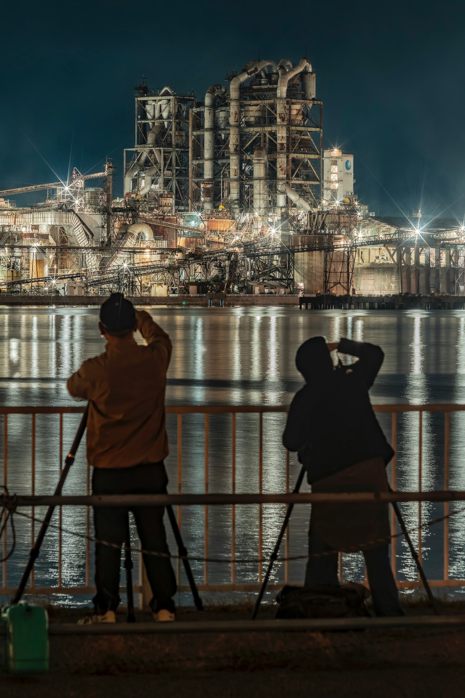 夜の工場の写真を撮る二人の人物と水面に映る工場の明かり