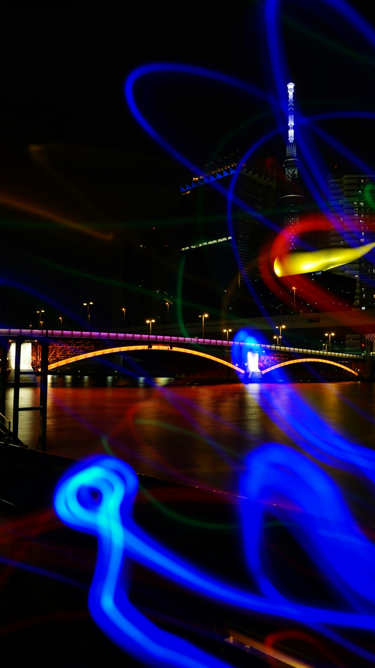 Bunte Lichtspuren über einer Brücke und Reflexionen im Wasser in einer nächtlichen Stadtlandschaft