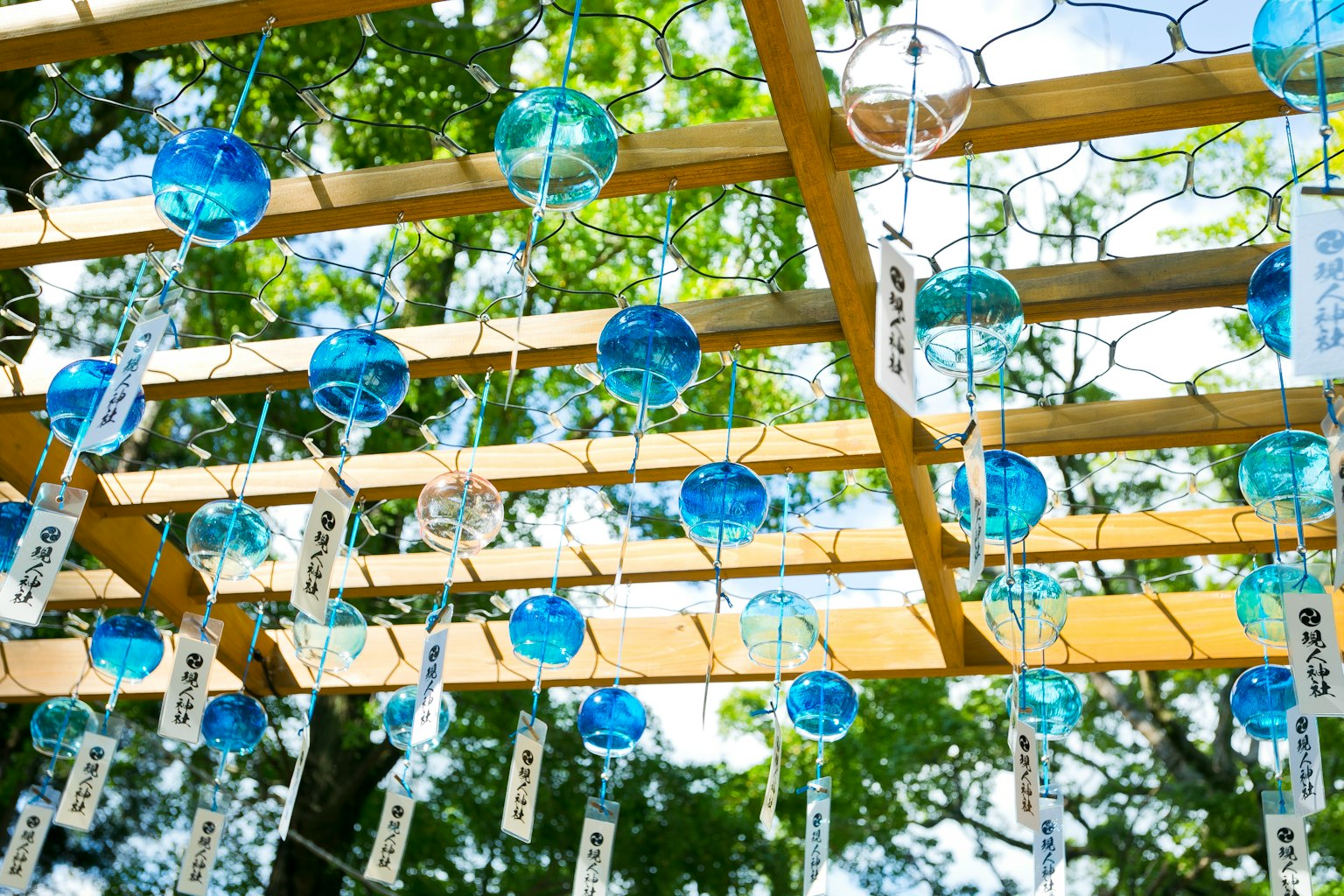 Blaue Windspiele hängen von einer Holzstruktur umgeben von grünen Bäumen
