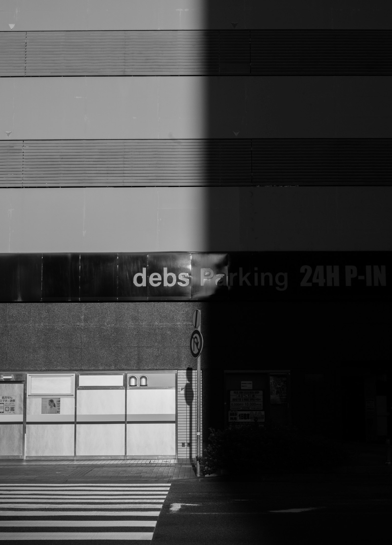 ภาพขาวดำที่มีความคอนทราสต์สูงของภายนอกอาคารพร้อมป้าย debs parking 24H P-IN