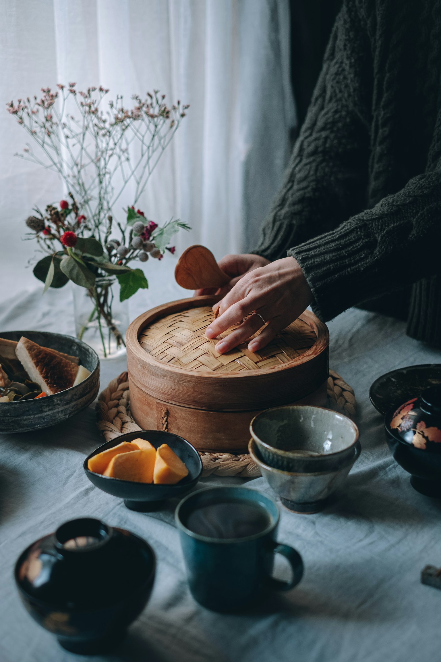 Hands opening a wooden steamer on a table surrounded by dishes and flowers