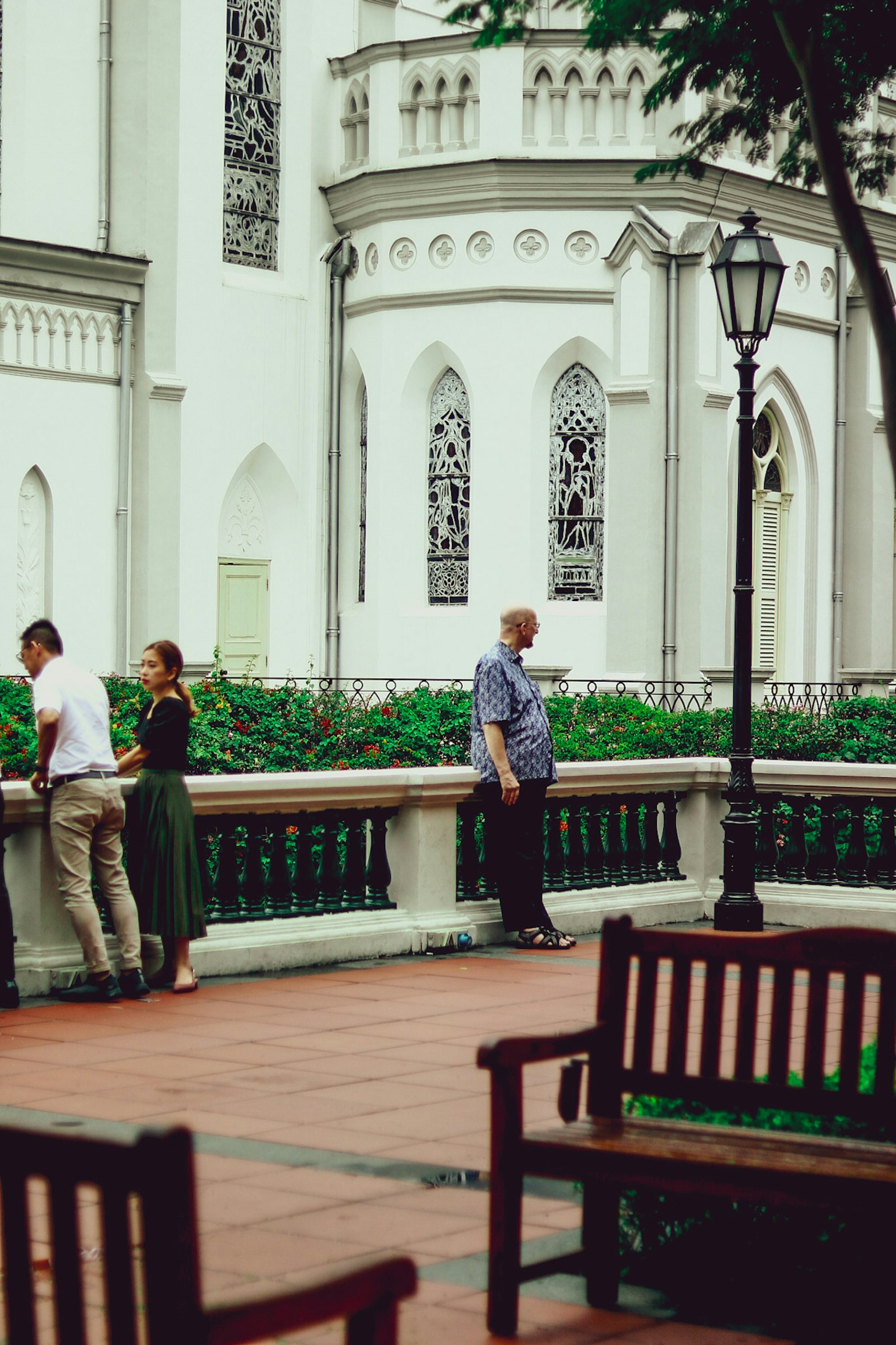 People standing by a white building with decorative windows and benches in a park