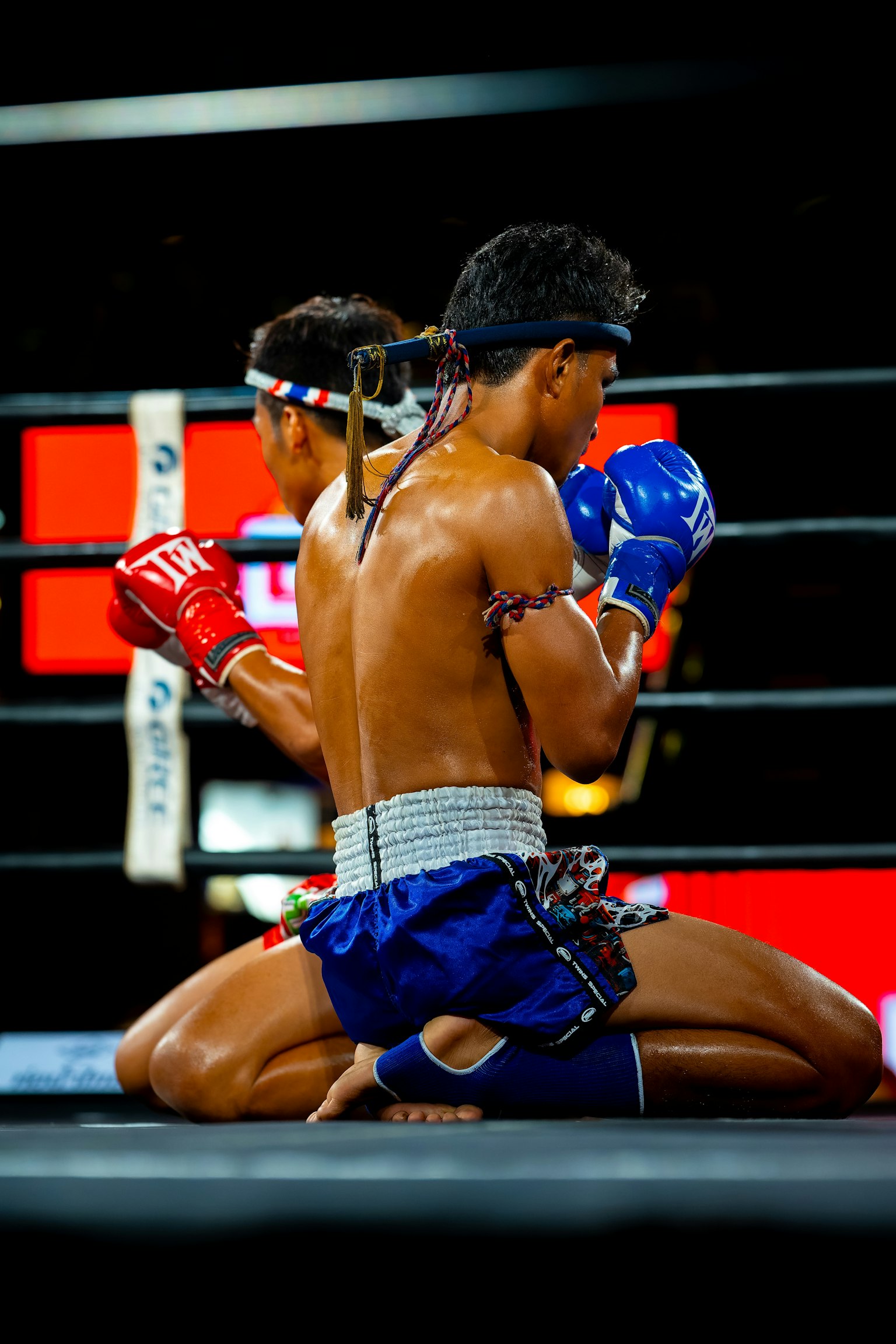 Hai võ sĩ Muay Thai quỳ gối ở giữa sàn đấu đeo găng tay đỏ và xanh với trang trí truyền thống