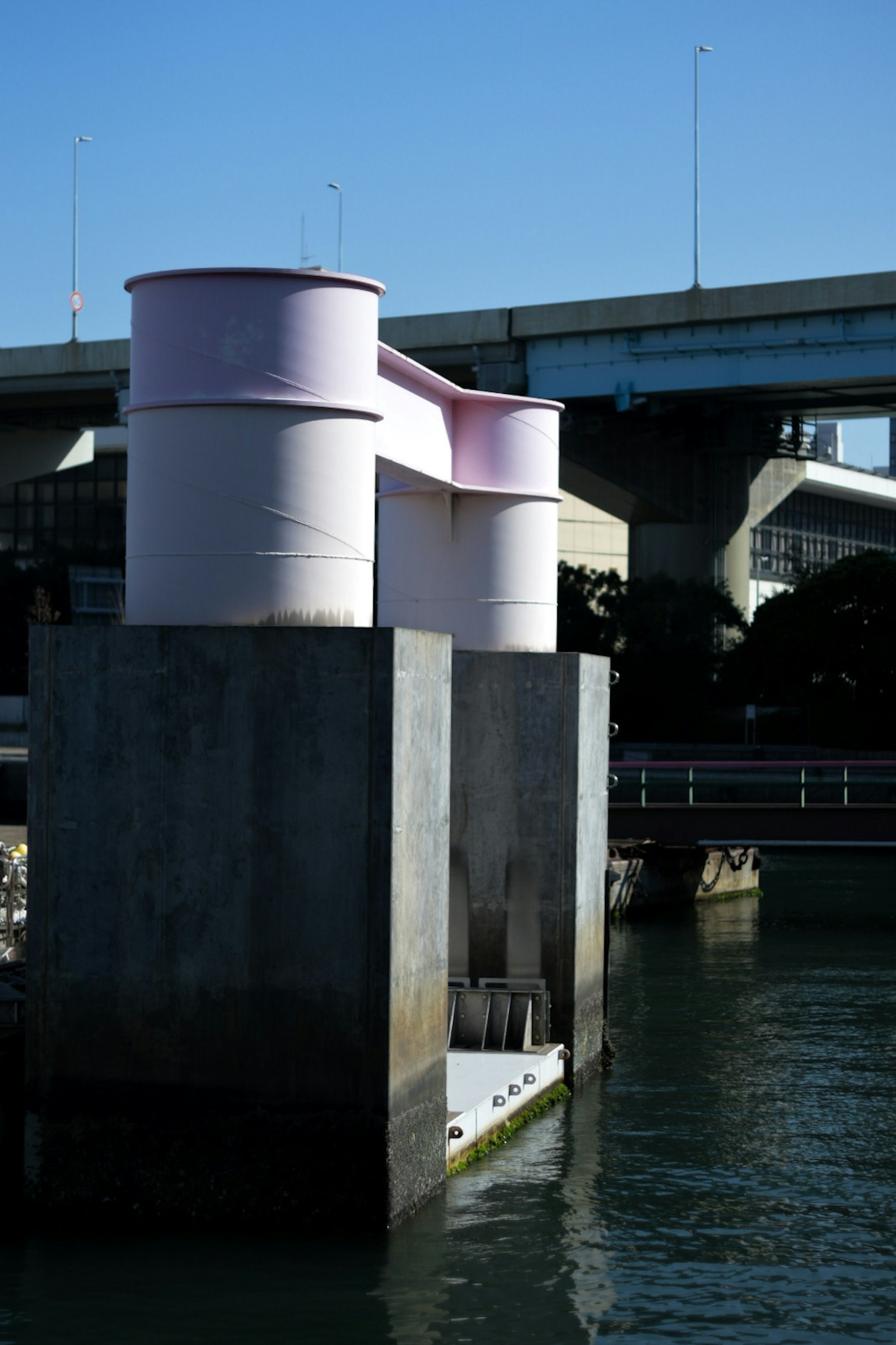 水辺に立つピンクのタンクとコンクリートの基礎 橋が背景に見える