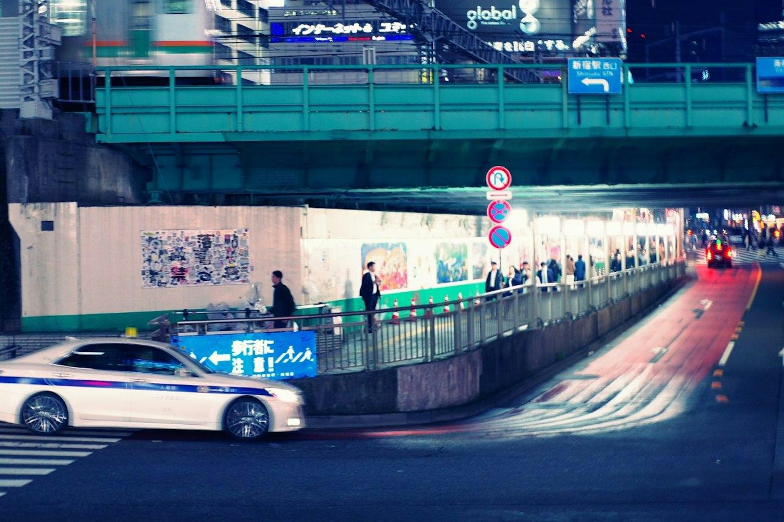 夜間城市場景，白色警車在藍色橋下行駛，人行道上有行人