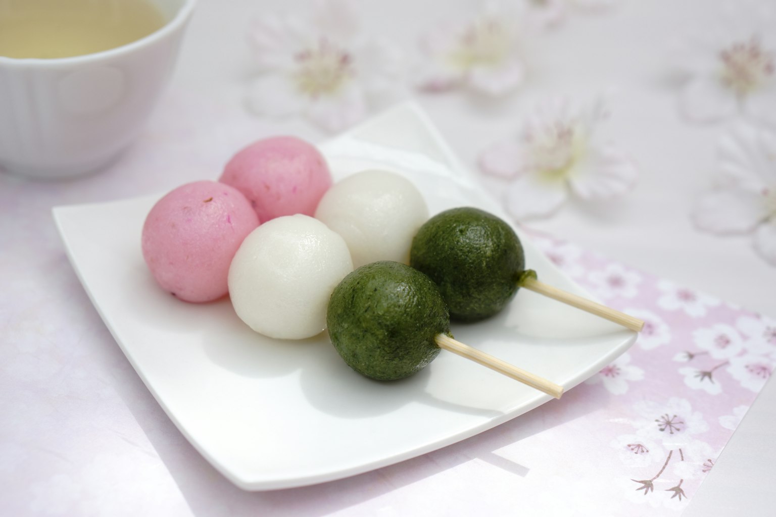Tres brochetas de dango en rosa, blanco y verde en un plato blanco con un fondo floral