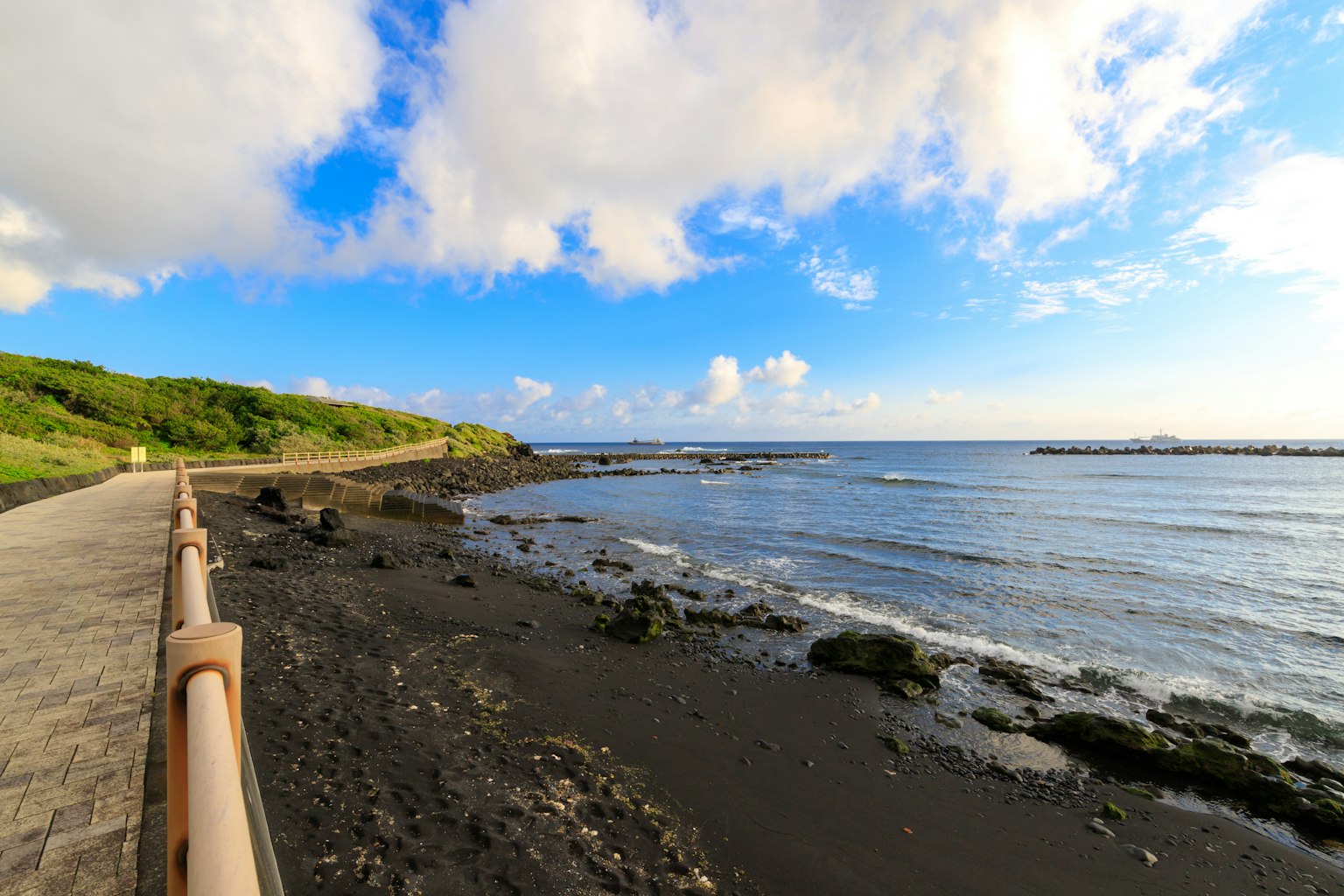 Coastal walkway with black sand and waves under a blue sky