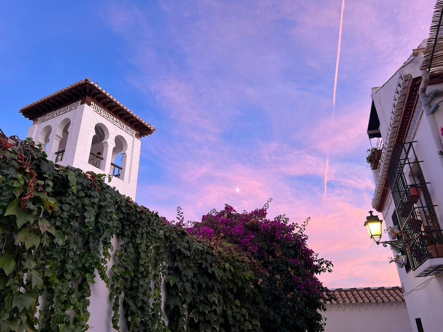 Vista escénica de un cielo al atardecer con tonos rosados que presenta un edificio de pared blanca y una torre adornada con enredaderas
