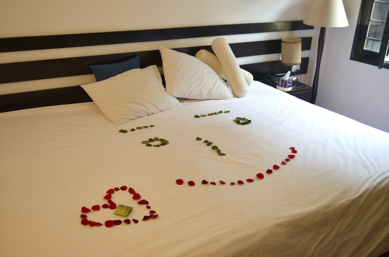 Una cama con sábanas blancas y almohadas decoradas con pétalos de rosa formando una cara sonriente y un corazón