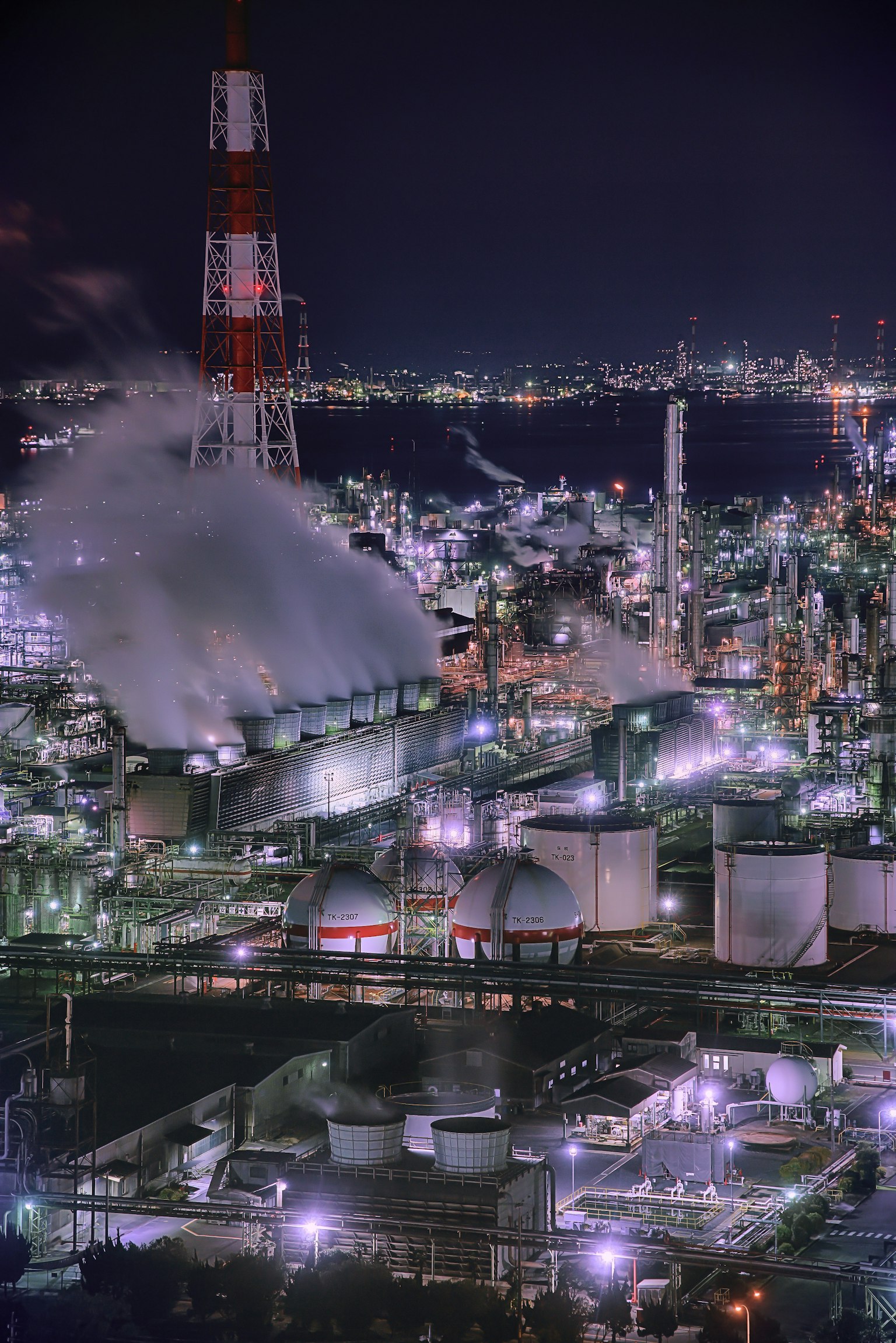 夜の工業地帯で輝くタンクと煙を上げる煙突の風景