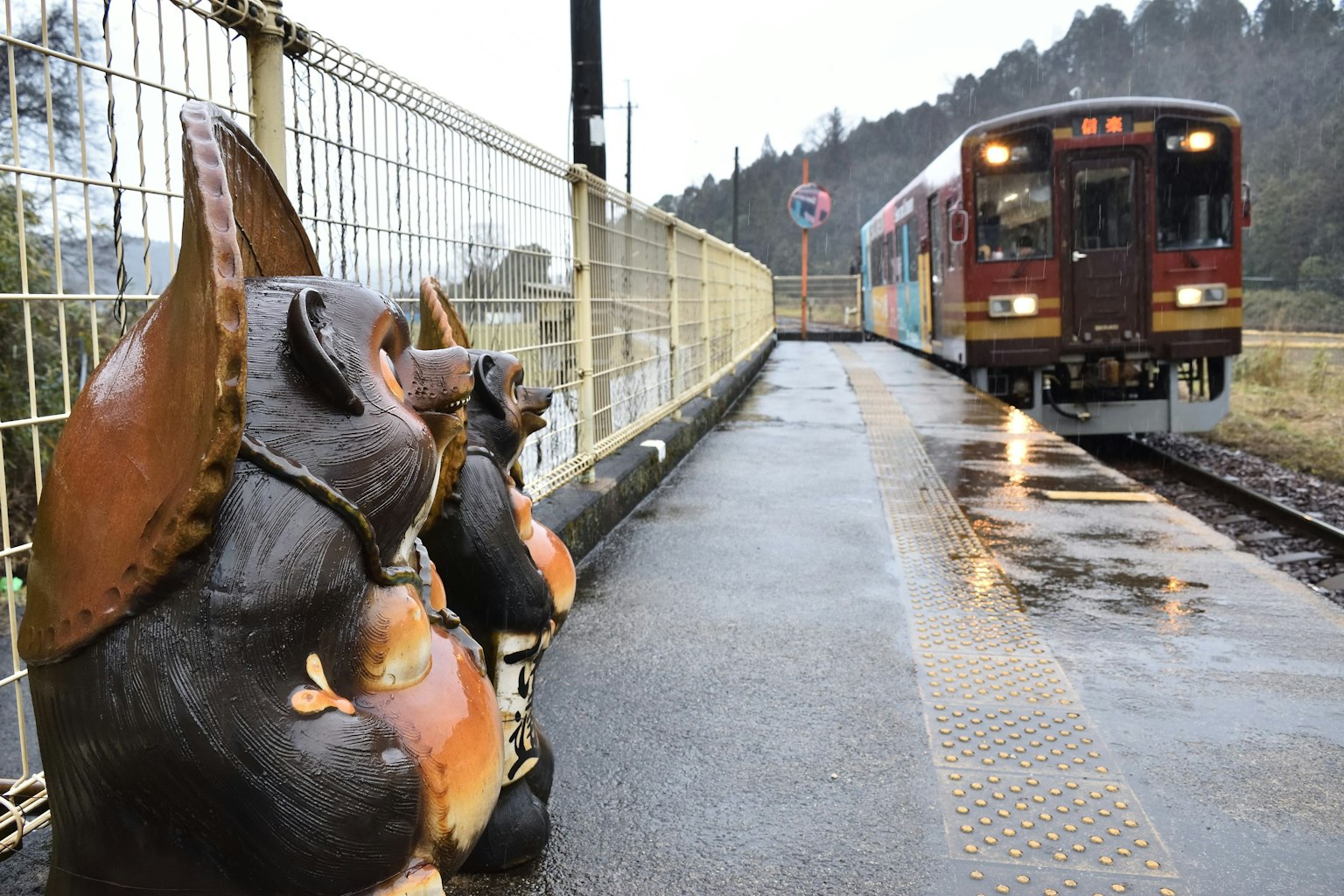 Pemandangan patung tanuki di stasiun kereta dengan kereta yang mendekat di bawah hujan