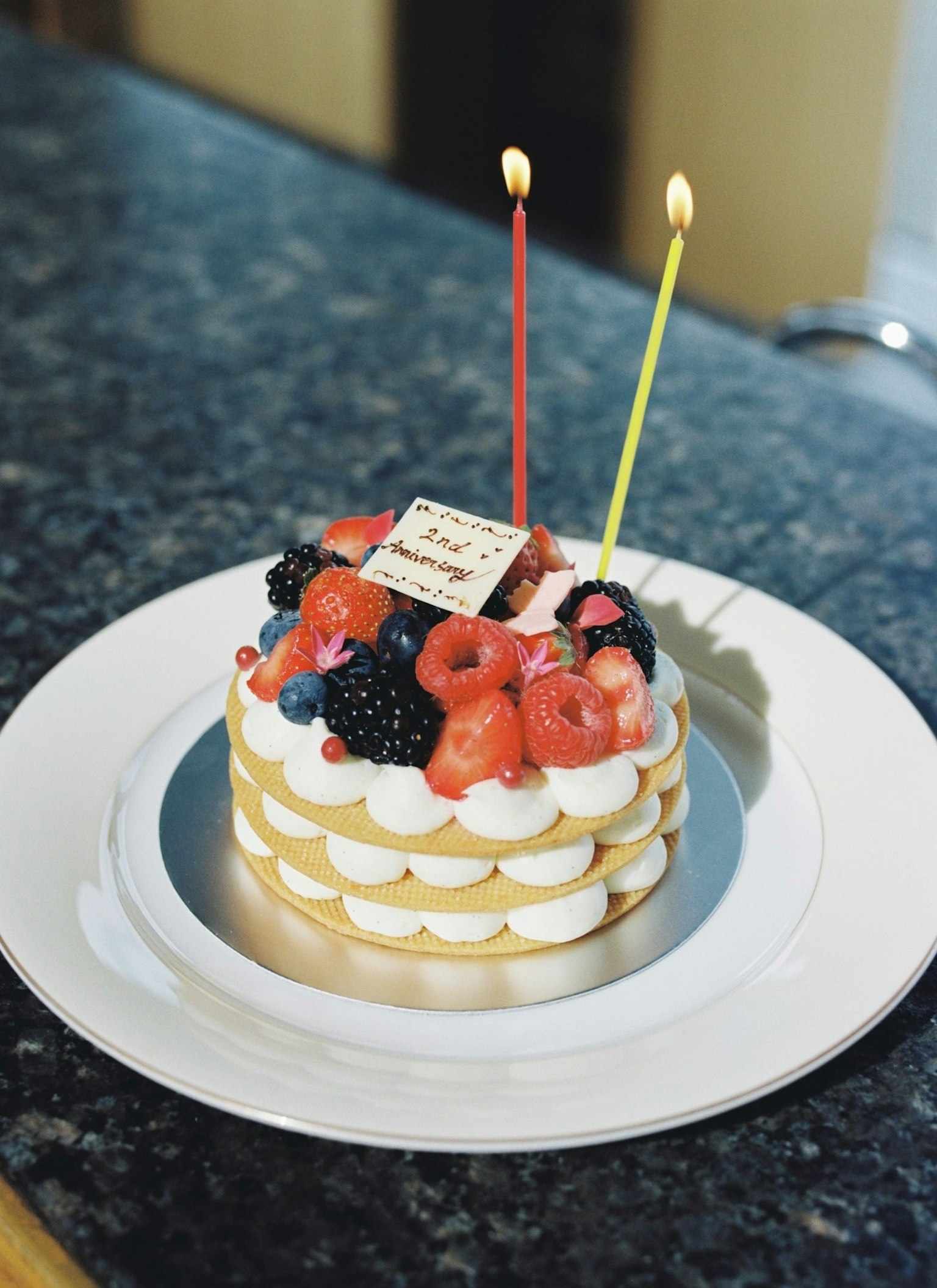 Gâteau d'anniversaire garni de crème et de fruits variés avec deux bougies et une plaque de message