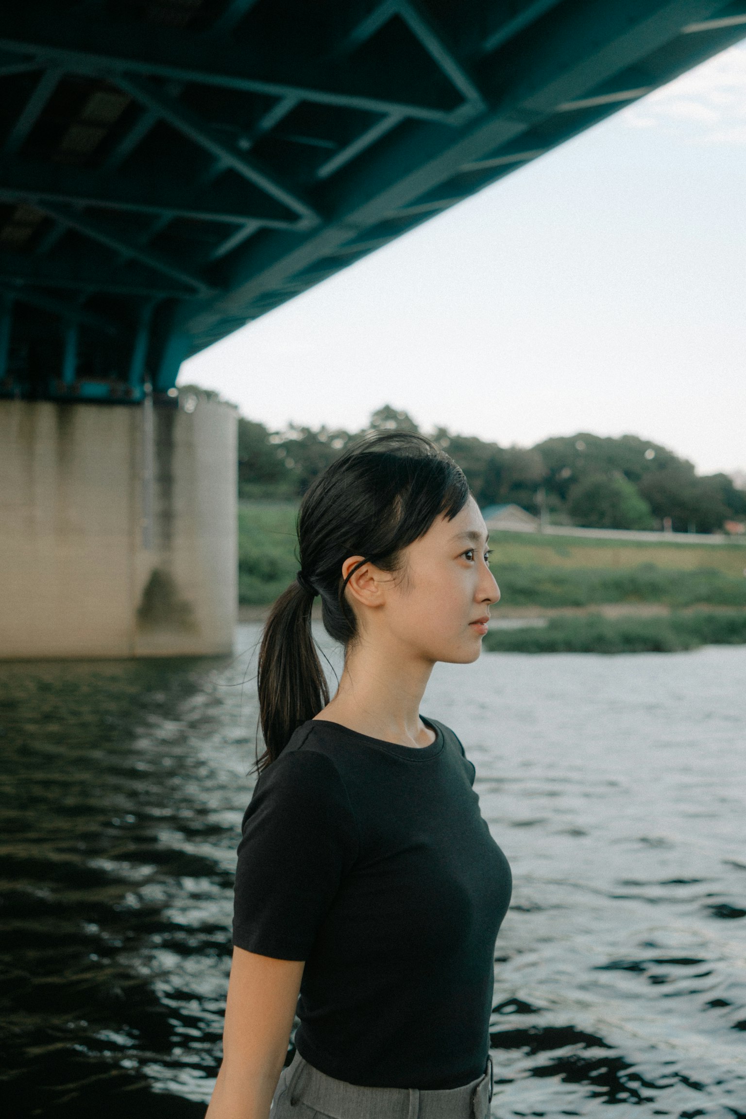 Profile of a woman standing by the water under a blue bridge
