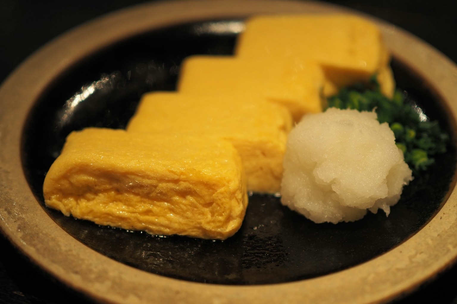 Tamagoyaki lezat disajikan di piring hitam dengan daikon parut dan bawang hijau