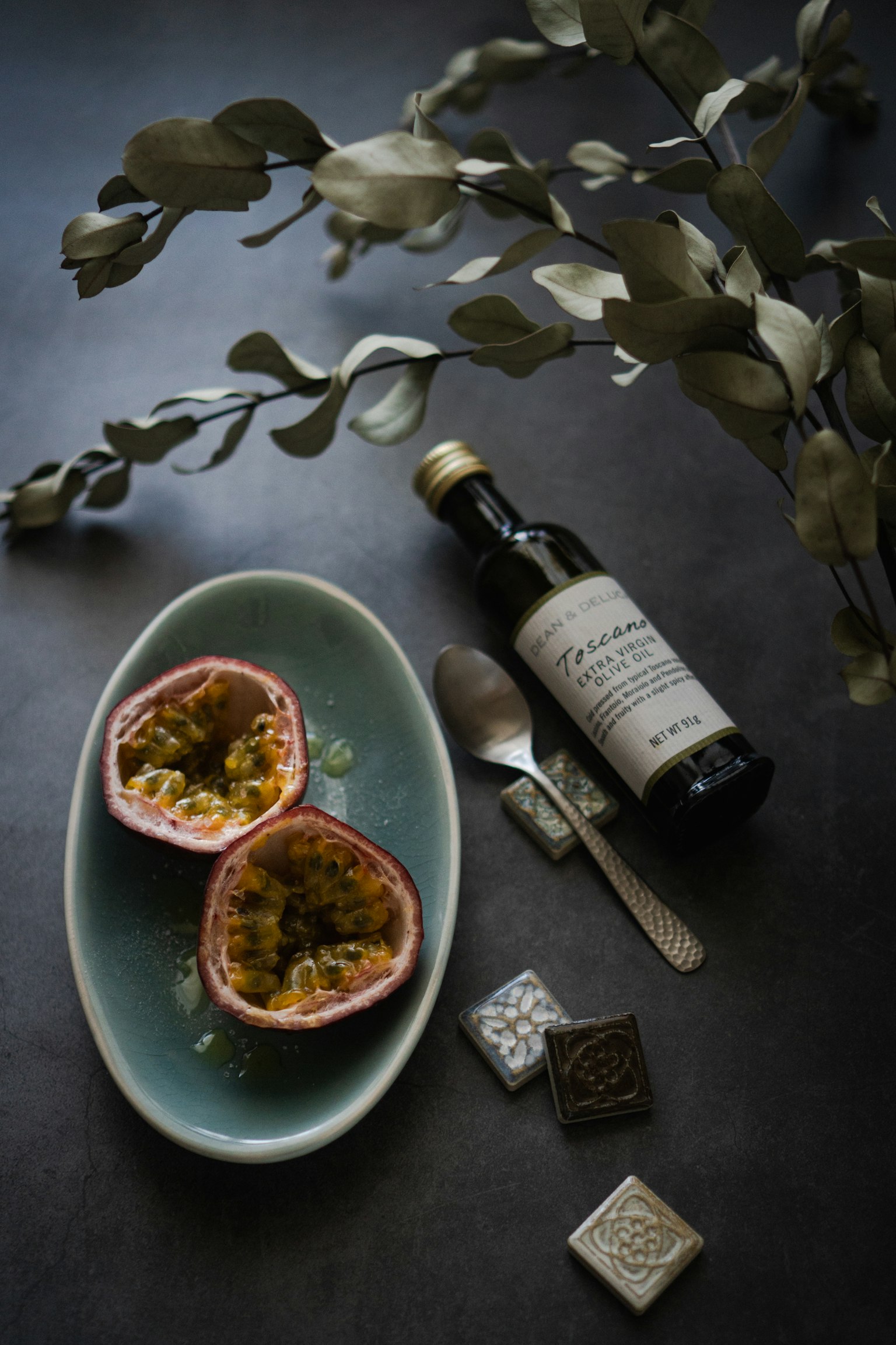 Fruits de la passion coupés en deux sur une assiette bleue avec une cuillère et une bouteille d'huile à côté de feuilles décoratives