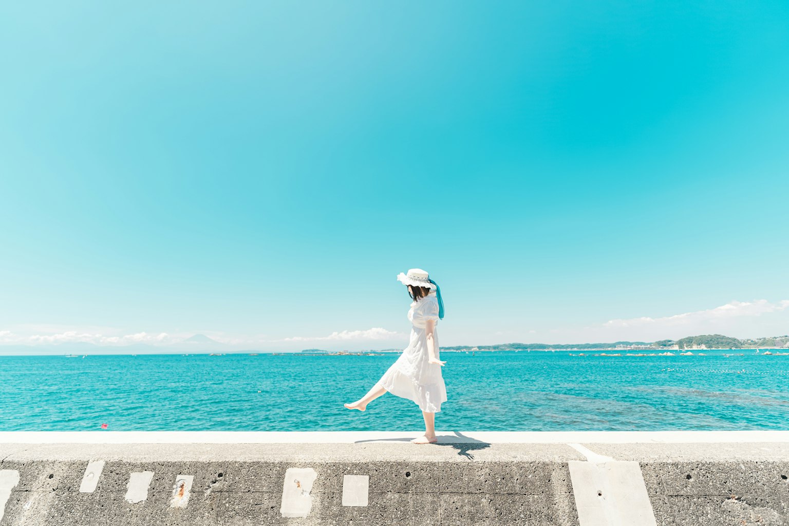 A woman in a white dress walking along a seawall under a blue sky
