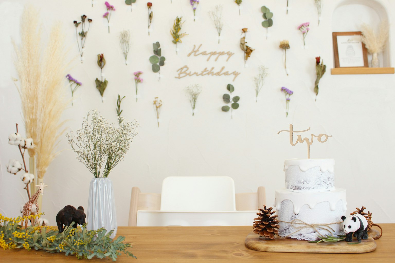 A birthday-themed table setting with a decorated wall featuring dried flowers and a cake