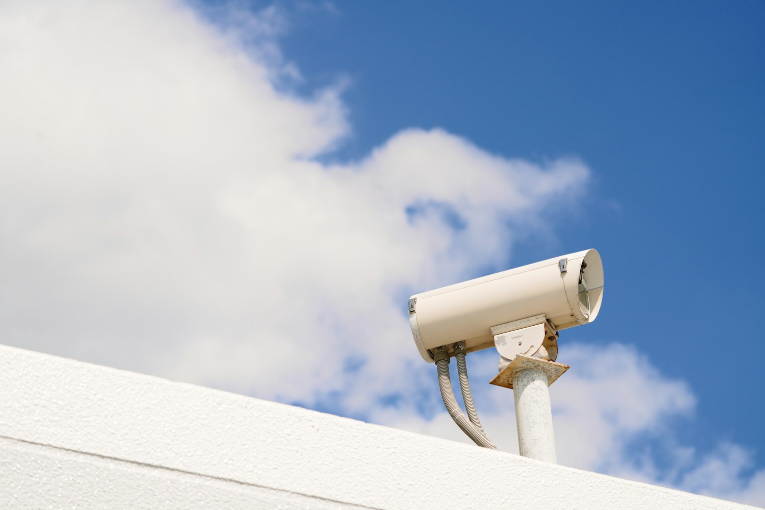 A white surveillance camera mounted on a wall under a blue sky with white clouds