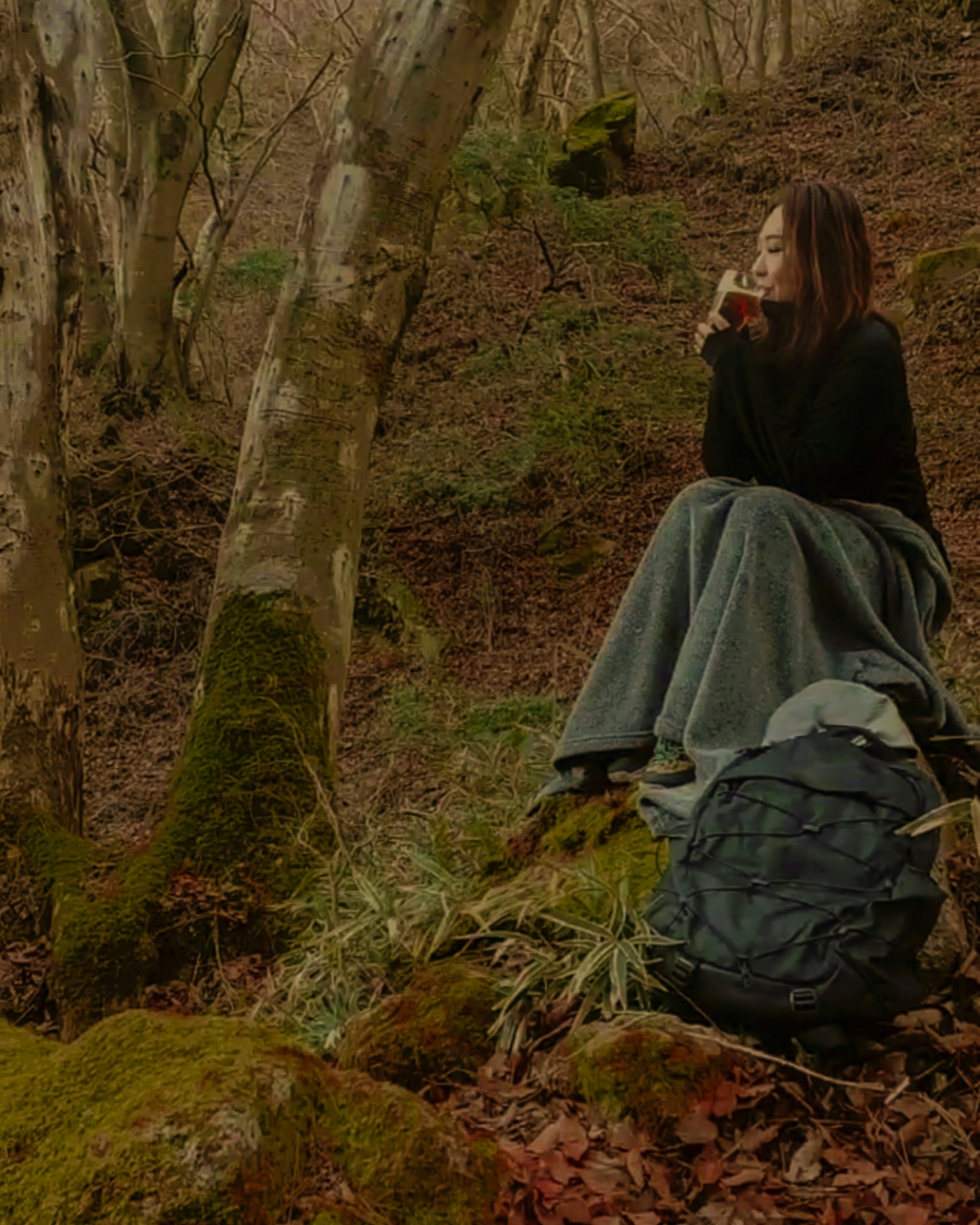 A woman sitting alone in a forest wearing warm clothing with a backpack beside her