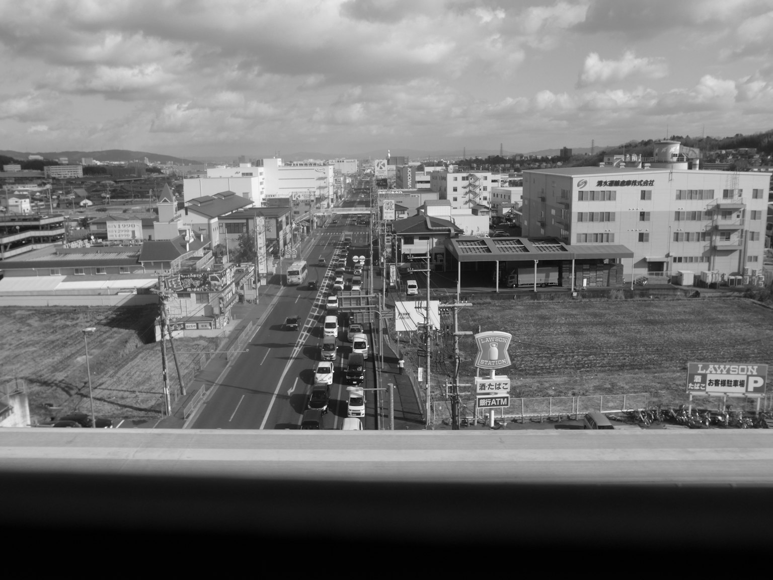 Paysage urbain en noir et blanc avec des véhicules alignés sur la route et des bâtiments visibles