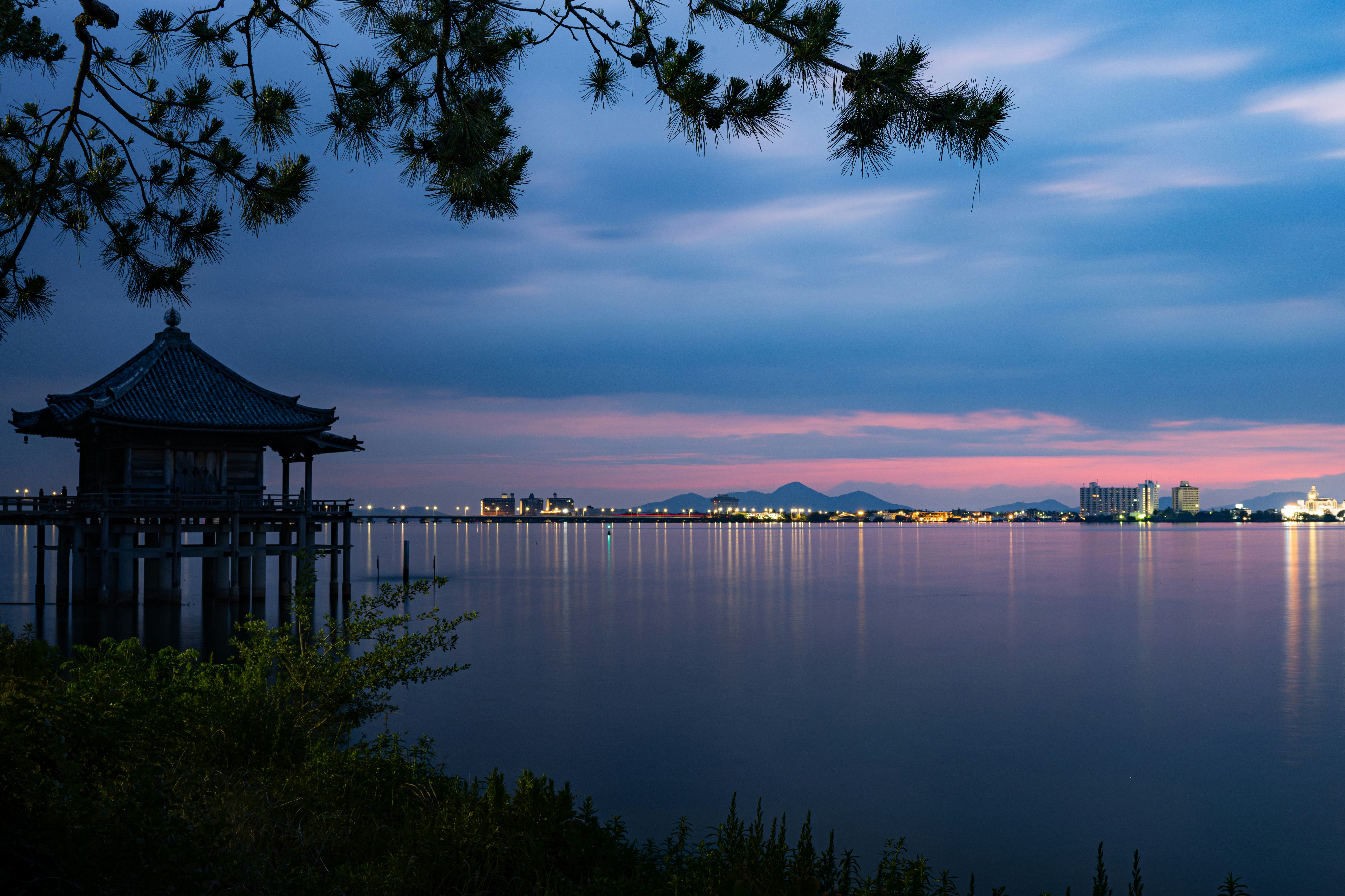 水面に映る夜景と月明かりの空、遠景の山並みと灯りのある海辺の風景