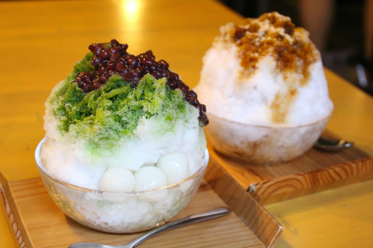 Two bowls of shaved ice on a wooden tray with green shaved ice and red bean topping