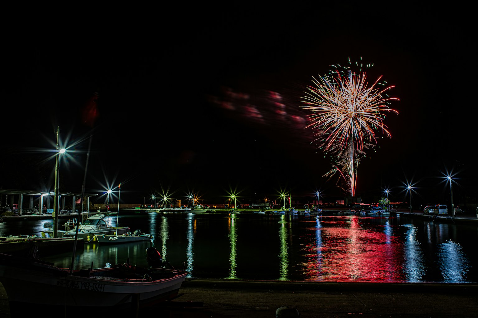 Fuochi d'artificio esplodono sopra un porto di notte con barche e luci riflesse nell'acqua