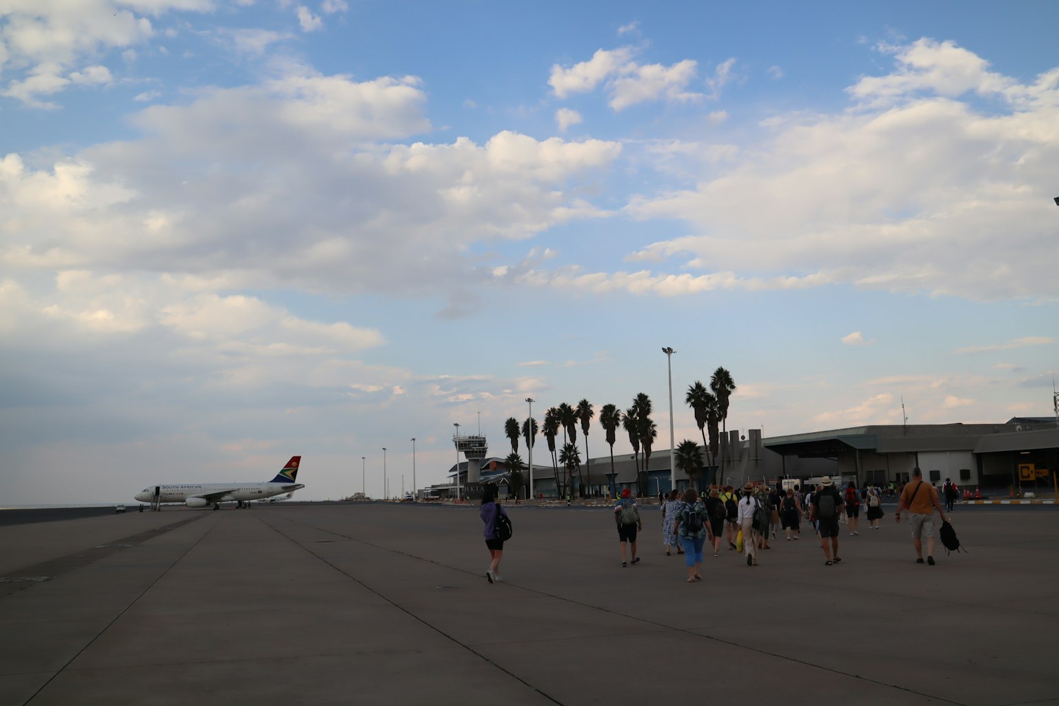 People walking on an airport runway with palm trees and a plane in the background