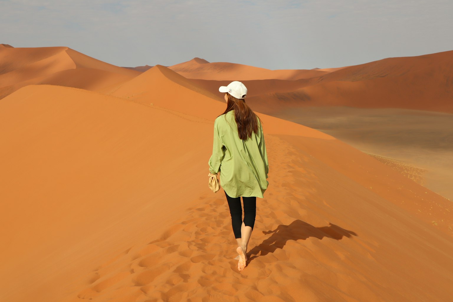 Mujer caminando sobre dunas de arena con una camisa verde y una gorra blanca