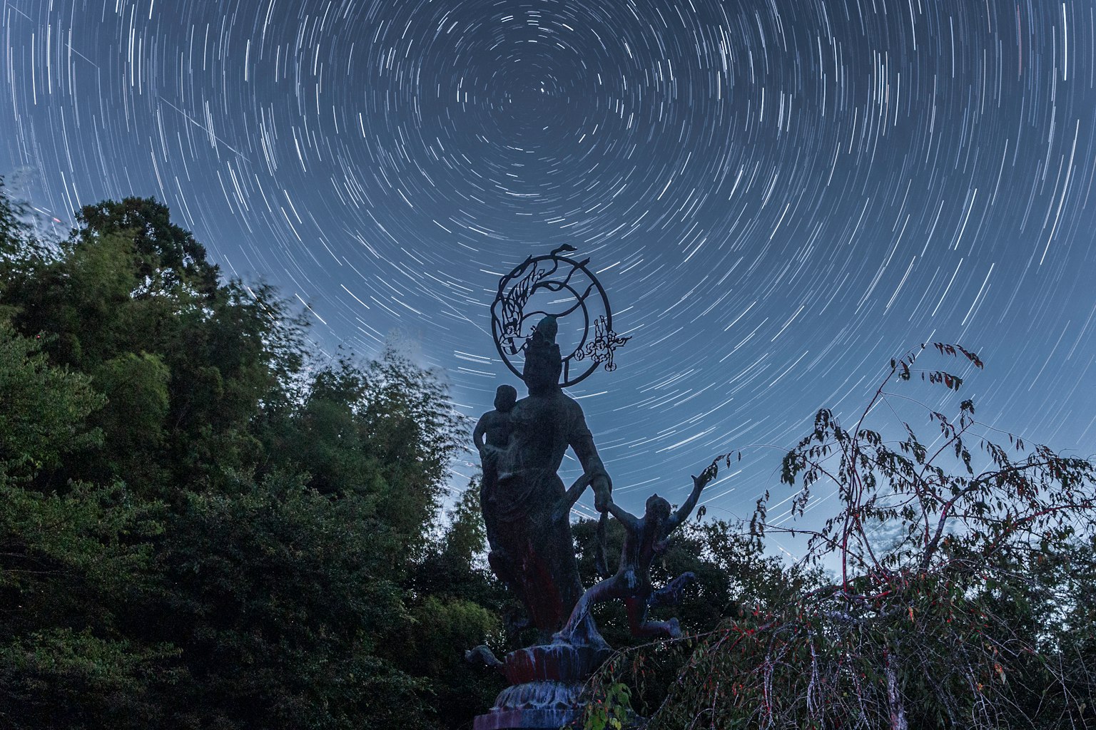 Gran estatua bajo un cielo estrellado con trazas de estrellas y siluetas de personas