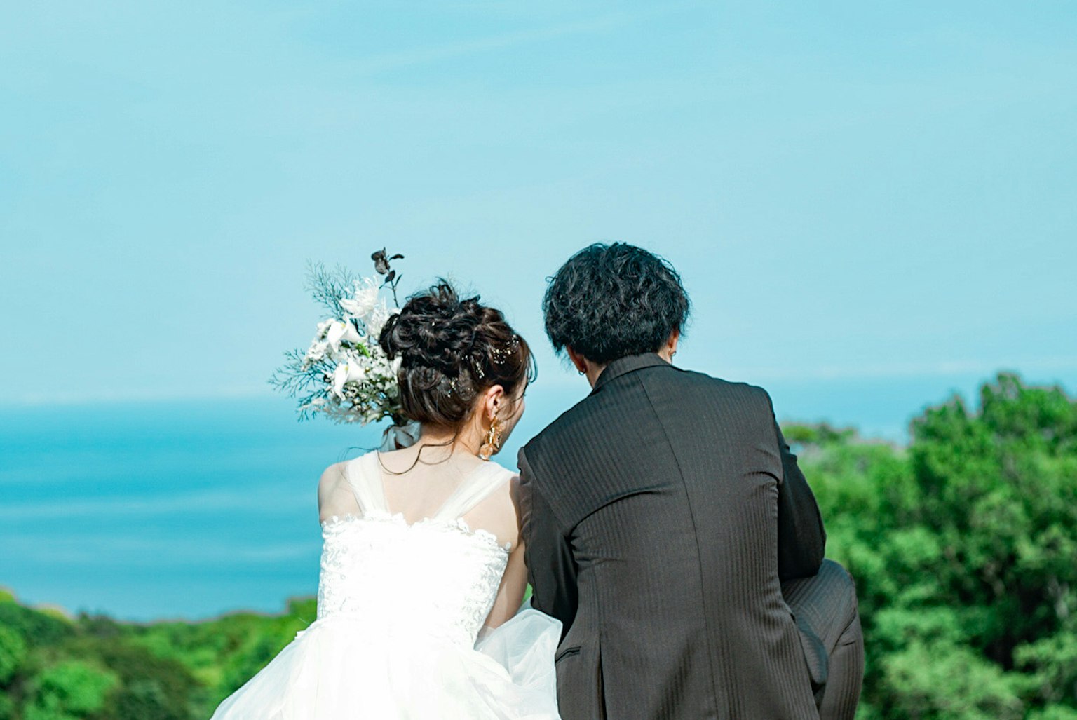Couple in wedding attire sitting together with ocean view and greenery