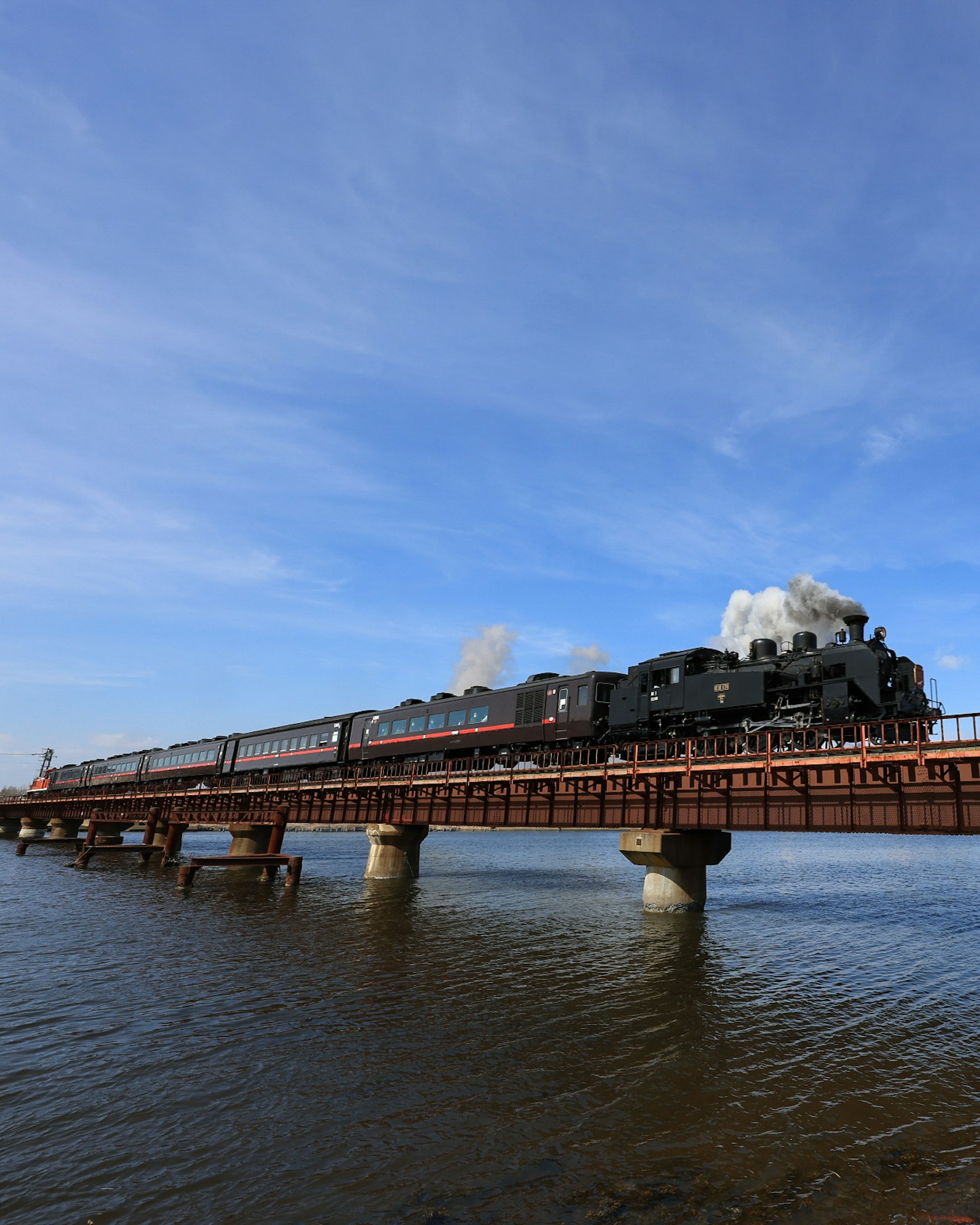 Una locomotiva a vapore che attraversa un ponte su acqua Riflessioni del treno sulla superficie Cielo blu con nuvole