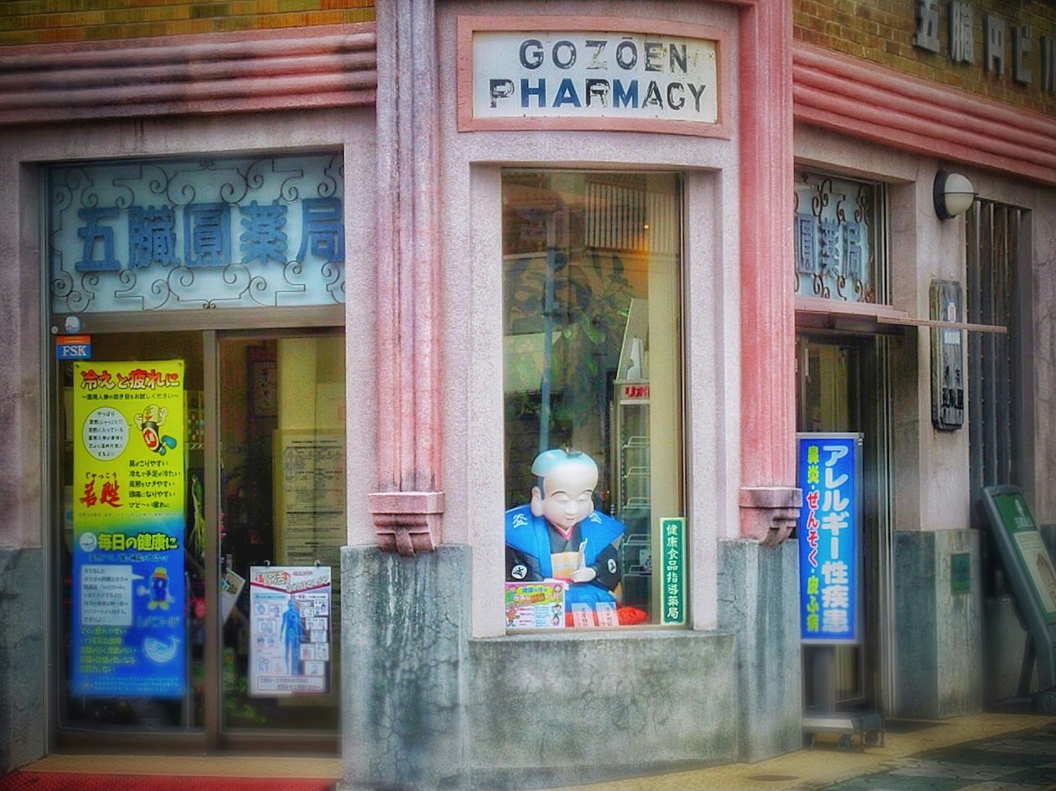 핑크색 외관의 약국 가게 앞에 서 있는 흰머리 약사 인형