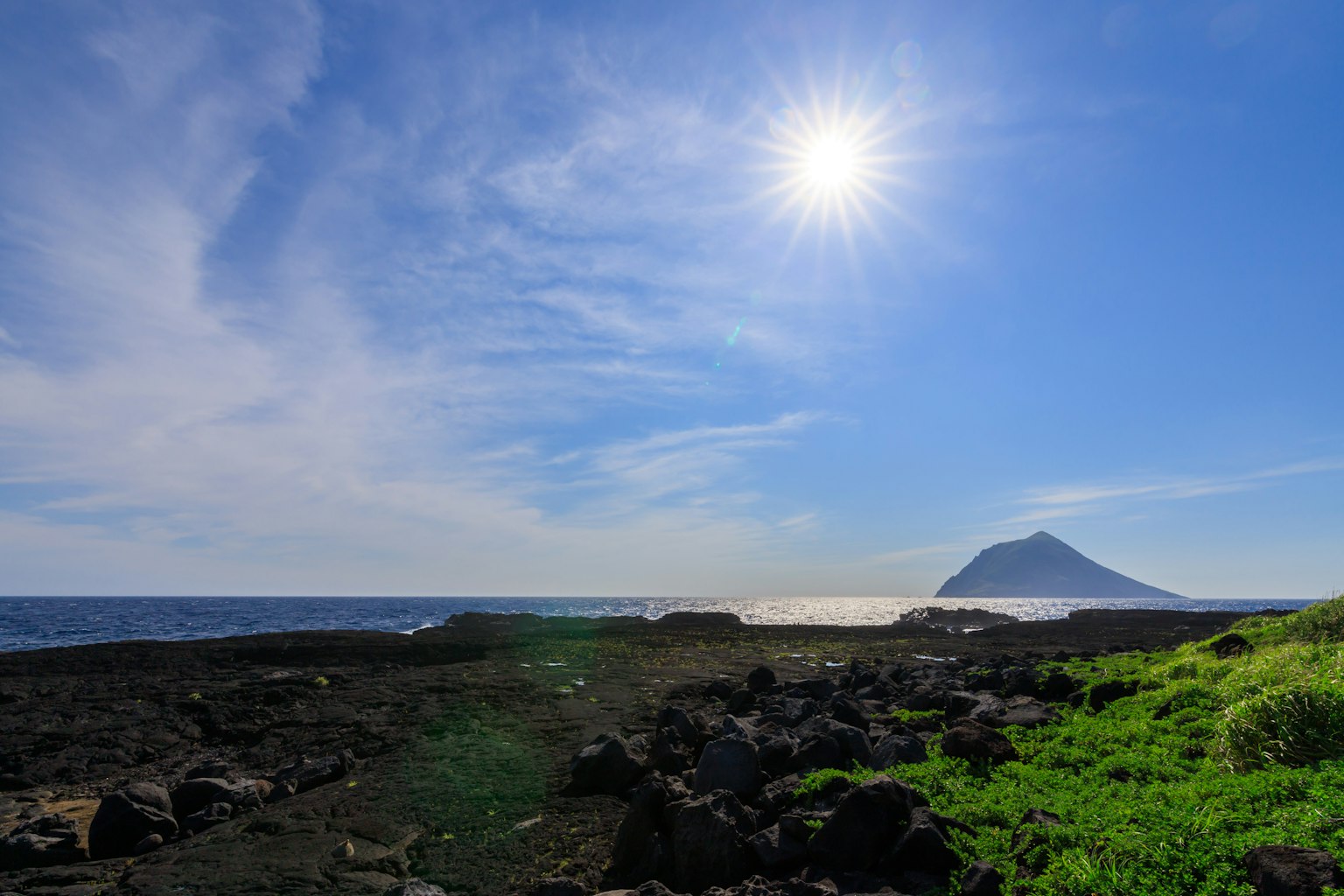 Coastal landscape with bright blue sky and sun black rocks and green grass prominent distant mountain