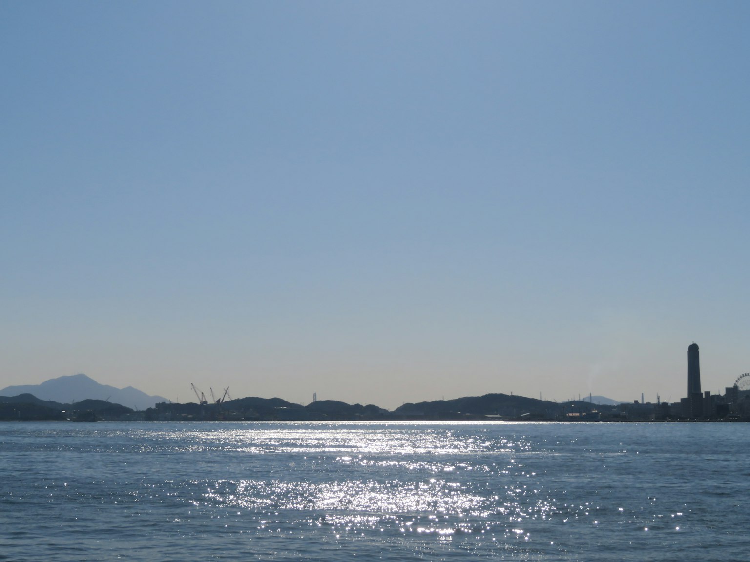 Clear blue sky over calm water with sunlit reflections, distant islands and a smokestack visible